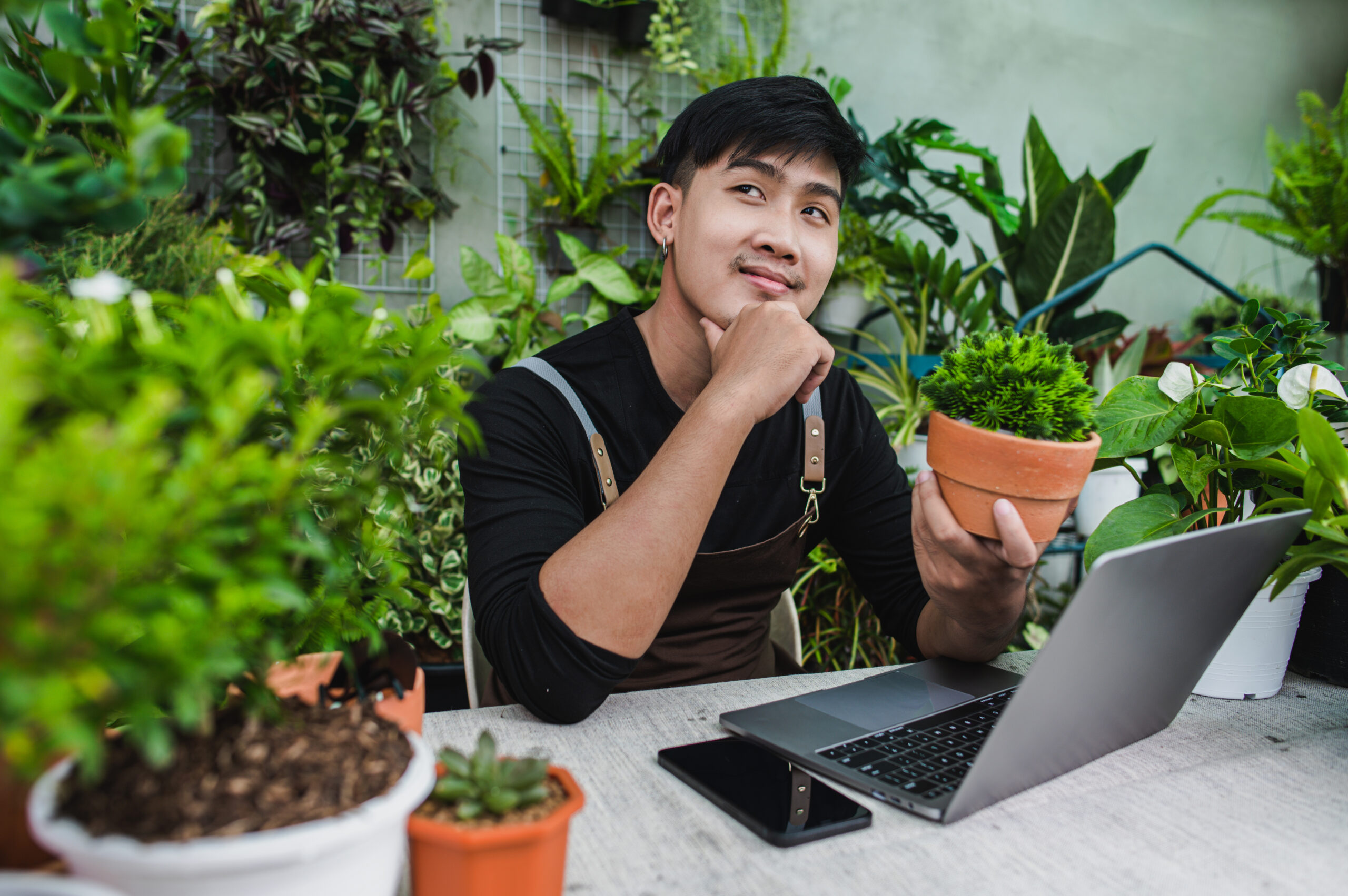 Happily gardener man use smartphone and laptop computer while online tutorial on potted plans in shop, vlogging or small business houseplant concept
